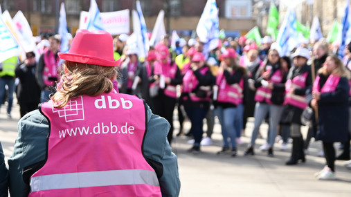 Das Foto zeigt eine Warnstreik-Demo des dbb. Im Vordergrund steht eine Frau, auf dem Rücken ihrer Weste steht das dbb-Logo. Im Hintergrund stehen weitere Demonstrierende. 