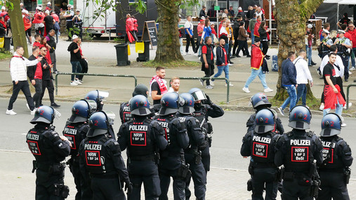 Foto zum Thema "Polizei Einsatzversorgung": Zu sehen sind Polizist*innen in Schutzkleidung, die Fans beobachten.