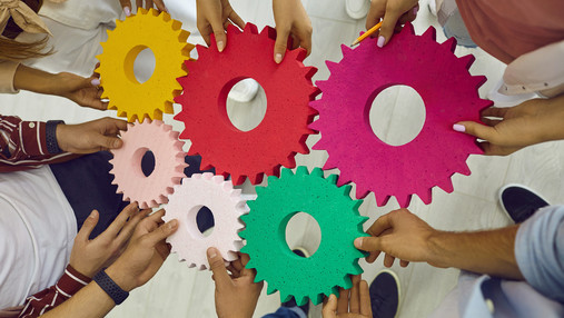 Foto zum Thema "Zukunft der Arbeit": Zu sehen ist ein Symbolbild. Personen, von denen man nur die Hände sieht, halten Modelle von insgesamt sechs Zahnrädern, die ineinandergreifen.