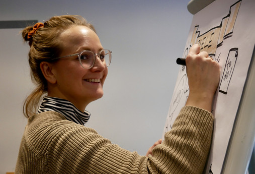 Zu sehen ist die Nahaufnahme einer Seminarteilnehmerin, die in die Kamera lächelt. In der Hand hält sie einen Edding, mit dem sie ihr Flipchart gestaltet.