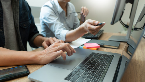 Foto zum Thema "was bedeutet agiles Arbeiten": Zu sehen ist ein Laptop und zwei Entwickler, nur ihre Hände und Oberkörper sind zu sehen. Es wird ersichtlich, dass sie gerade über ein Projekt sprechen.
