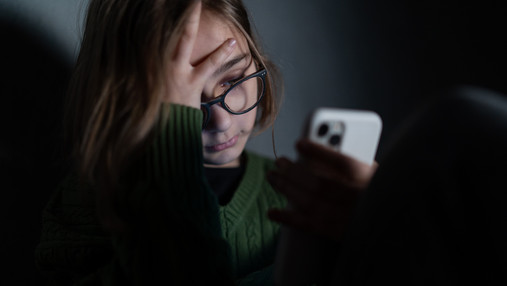 Foto zum Thema "Kinder- und Jugendtelefon": Zu sehen ist ein Mädchen, das in der Dunkelheit sitzt und verzweifelt aufs Smartphone schaut.