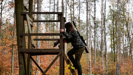Zu sehen ist eine Jägerin, die auf einen Hochsitz klettert.