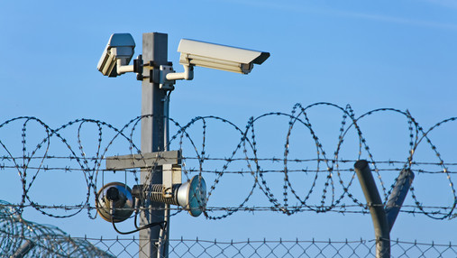 Foto zum Thema "BSBD": Zu sehen sind Sicherungsanlagen eines Gefängnisses. Kameras und Stacheldraht dominieren das Bild.