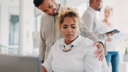 Foto zum Thema sexuelle Belästigung am Arbeitsplatz: Zu sehen ist eine Frau, die am Schreibtisch im Büro sitzt, und von einem Mann an der Schulter berührt wird. Ihr Blick verrät, dass es ihr nicht gefällt.