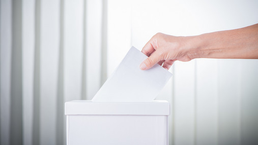 Foto zum Thema "einmal Wahlhelfer, immer Wahlhelfer": Zu sehen ist eine Wahlurne und die Hand einer Person, die gerade ihre Stimme einwirft.