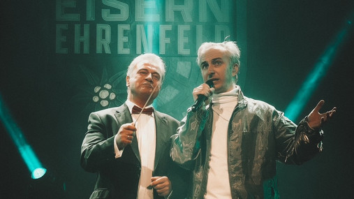 Foto zum Thema "Claus Weselsky und Jan Böhmermann": Zu sehen sind Weselsky und Böhmermann auf der Bühne der Jahrhunderthalle in Frankfurt. Weselsky hat einen Taktstock in der Hand, Böhmermann ein Mikrofon.