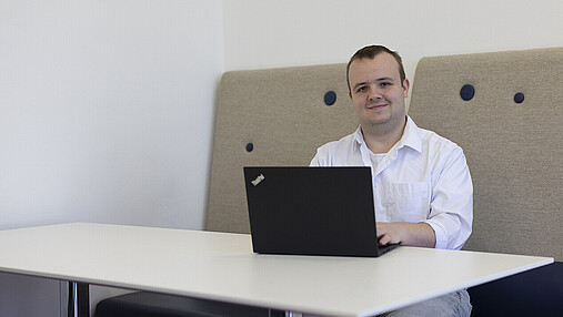Foto zum Thema "Fachinformatiker*in für Systemintegration": Zu sehen ist Alexander Bacher, Fachinformatiker für Systemintegration beim Deutschen Patent- und Markenamt. Er sitzt mit einem Laptop an einem Tisch.